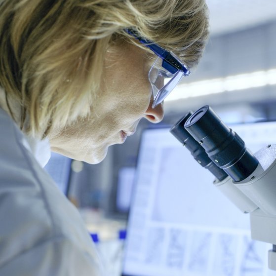 Forschung Weibliche Person im weißen Laborkittel und Schutzbrille schaut durch ein Mikroskop. Im Hintergund ein Computerbildschirm.