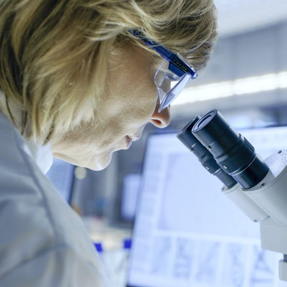 Forschung Weibliche Person im weißen Laborkittel und Schutzbrille schaut durch ein Mikroskop. Im Hintergund ein Computerbildschirm.