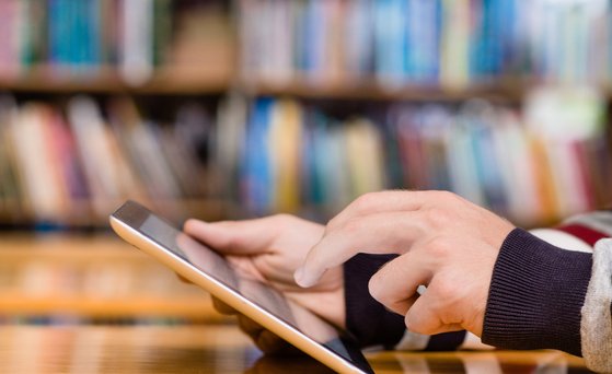 Tablet wird bedient, Hände im Close-Up, Bücherwand im Hintergrund unscharf
