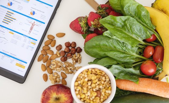 Von oben fotografiertes gesundes Essen, Nüsse, Bannanen, Erdbeeren, Salat, Tomaten liegen auf einer weiße, Untergrund, daneben ein Tablett auf dessen Display  Diagramme, Zahlen abgebildet sind. 