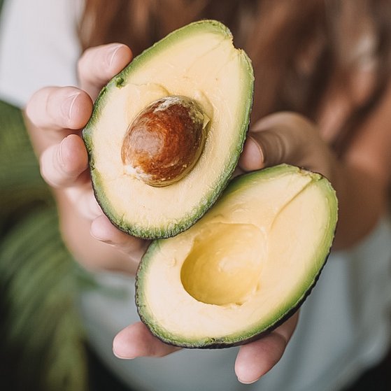 Ernährungsrisiken Frau hält aufgeschnittene Avocado in die Kamera. In einer Hälfte befindet sich noch der Kern.