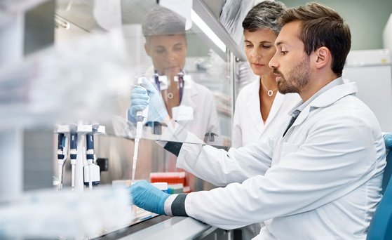 Zwei Wissenschaftler sind im Labor. Einer von ihnen pipettiert hinter einem Schutzglas. Wissenschaftlerin beobachtet ihn dabei.