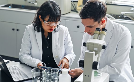 Zwei Menschen unterhalten sich und schauen auf ein Tablet. Ein Mikroskop und ein Laptop sowie Bechergläser stehen auf dem Tisch. Beide tragen Laborkittel.