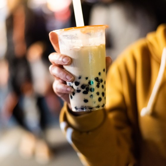 Plastikbecher mit Bubble Tea wird in der Hand gehalten. Person trägt senfgelben Kaputzenpullover, Kopf nicht sichtbar, Hintergund unscharfe Menschen