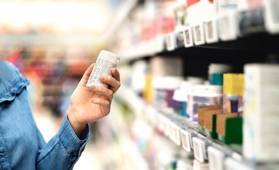 Hand hält Arzneimittelverpackung mit Etikettentext über Inhaltsstoffe in den Händen. Im Hintergrund sieht man unscharf die Regale eines Drogeriemarktes.