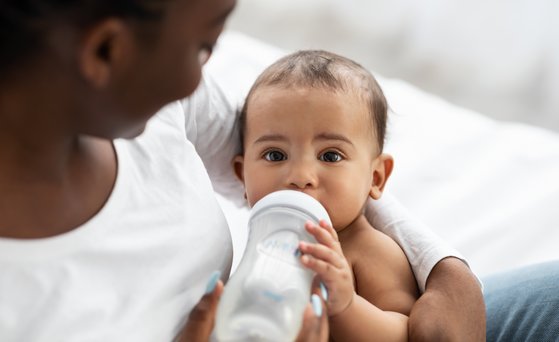 Baby im Arm der Mutter mit Trinkflasche. Blickt direkt in die Kamera.