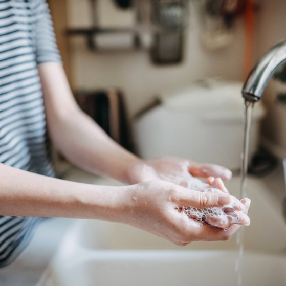 Person mit geringeltem T-Shirt im Anschnitt der linken Bildhälfte, die ihre Hände in einem Waschbecken mit Seife wäscht.