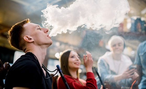 A group of young people are smoking a shisha, a man in the foreground blows smoke out.