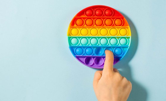 Rundes Fidget Toy in Regenbogenfarben liegt auf hellblauer Fläche. Ein Finger drückt Silikon-Bubble nach innen.