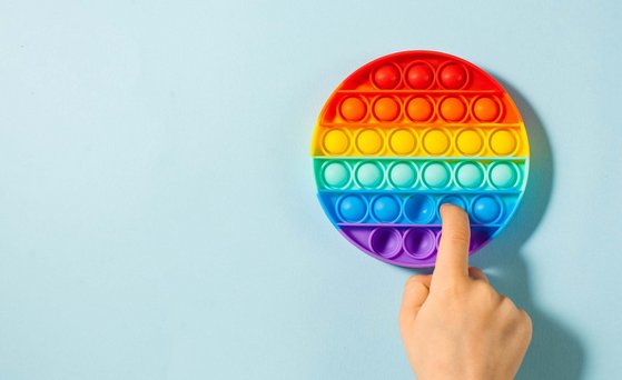 Round fidget toy in rainbow colours lies on a light blue surface, hand in the picture from below, one finger pushes the silicone bubble inwards