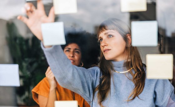 Zwei Frauen stehen hinter einer Glasscheibe und schauen sich Post-it an, die an der Scheibe kleben.