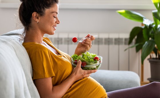 Schwangere Frau sitzt auf dem Sofa mit einer Schüssel Salat.