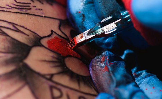 Nahaufnahme von einer Hand, die einen blauen medizinischen Handschuh trägt und rote Farbe mit der Tätowiermaschine in Haut einarbeitet.