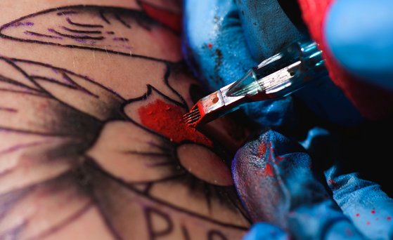 Tätowierfarbe Nahaufnahme von einer Hand, die einen blauen medizinischen Handschuh trägt und rote Farbe mit der Tätowiermaschine in Haut einarbeitet.